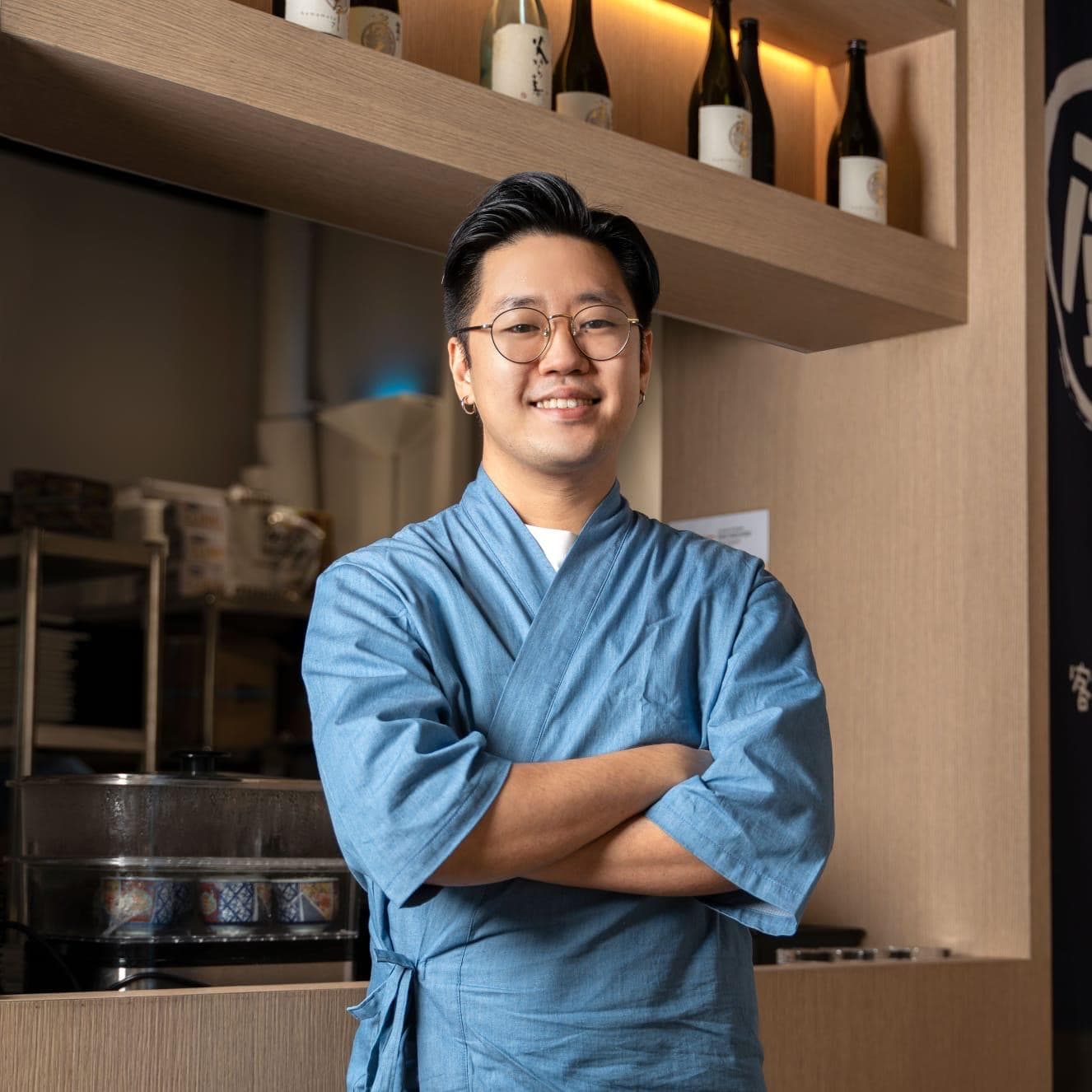 Dominic, arms crossed at the counter, sake bottles on the shelf behind him
